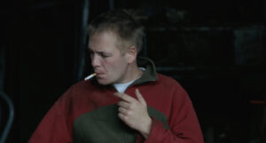 Filmstill aus Alex in den Feldern. Ein blonder Mann in enem roten Pullover raucht eine Zigarette und schaut zur Seite.
