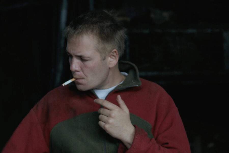 Filmstill aus Alex in den Feldern. Ein blonder Mann in enem roten Pullover raucht eine Zigarette und schaut zur Seite.