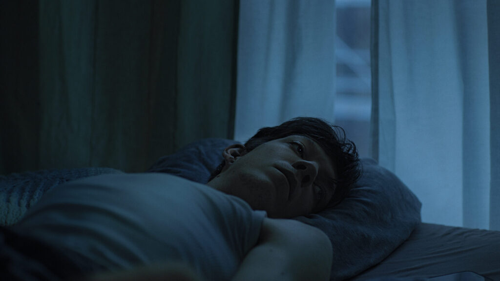 Filmstill aus Beben: ein junger Mann liegt im Halbdunkel vor einem Fenster in einem Bett.