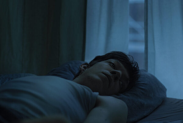 Filmstill aus Beben: ein junger Mann liegt im Halbdunkel vor einem Fenster in einem Bett.